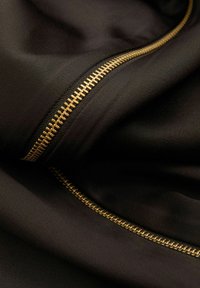 Schwarzer Stoff mit einer glatten Textur, der mit einem goldenen Reißverschlussdetail versehen ist. Der Reißverschluss hat sichtbare Zähne und verläuft geschwungen entlang des Randes des Materials.