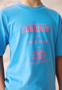 Tricou din bumbac albastru cu grafice imprimate roz, incluzând text și logo. Guler rotund și mâneci scurte, textură netedă.