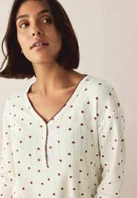 Valkoinen toppi punaisilla sydänkuvioilla, V-pääntie ja koristeellinen reunus. Valmistettu pehmeästä kankaasta, joka tuntuu teksturoidulta. Lyhyet hihat.