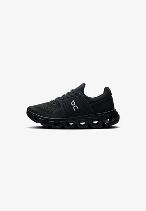 Schwarzer Turnschuh mit Mesh-Obermaterial, Schnürung und einer dicken, wellenförmigen Gummisohle. Verfügt über ein Logo an der Seite und strukturierte Designelemente.