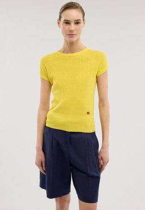 Kobieta ubrana w żółtą, teksturowaną bluzkę z krótkim rękawem i granatowe plisowane szorty do kolan, stojąca na jednolitym tle.