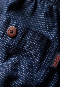 Tela azul marino con un patrón texturizado y pequeñas motas claras, que incluye un botón marrón oscuro con "SUPERDRY" y un acento de etiqueta roja.