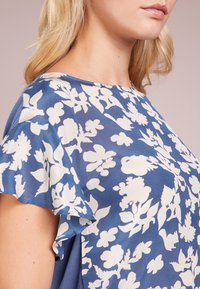 Top con estampado floral en azul y blanco, con mangas cortas con volantes, cuello redondo y una textura de tela suave y brillante.