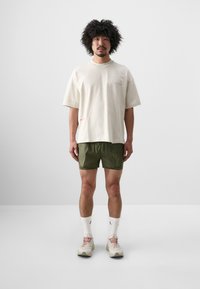 Beige bomulls t-shirt med lös passform, tillsammans med gröna shorts i lätt tyg. Skorna har en neutral färg med rosa detaljer.