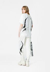 Camiseta blanca de gran tamaño con bordado de alas en negro y plateado en la parte posterior. Combinada con pantalones de chándal blancos, también adornados con detalles de alas.