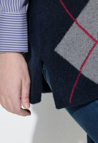 Ruka poblíž stehna, oblečená v modrých džínách, námořnický svetr s argyle vzorem v červené a šedé barvě, a rukáv košile s modro-bílými pruhy.