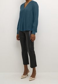 Blouse teal à manches longues et décolleté en V, associée à un pantalon cropped en faux cuir noir et des mules beiges à talons avec un détail en nœud.
