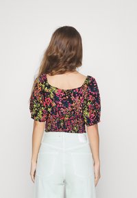 Černá květinová blůza s barevnými růžovými a žlutými květy, nabíranými rukávy a elastickým zadním designem. Stylizováno s kalhotami v světlých barvách.