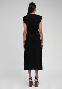 Robe noire sans manches, de longueur mi-mollet, avec une taille cintrée et une texture subtile. Le dos présente un détail en trou de serrure au niveau du cou.