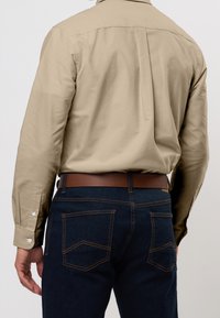 Beige langärmeliges Hemd mit geknöpften Manschetten, kombiniert mit dunklen Jeans aus Denim mit kontrastierenden Nähten und einem braunen Gürtel.
