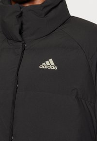 Svart puffjacka med hög krage, med en subtil Adidas-logga i ljusgrått, knäppning med knappar och textil med struktur.