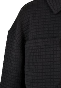 Manteau en tissu matelassé noir avec motif carré texturé, col visible et grande poche avant sur la partie supérieure de la manche.
