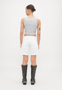Top cortado gris, pantalones cortos blancos y botas de cuero negras hasta la rodilla. Tatuaje visible en el brazo, fondo sencillo.