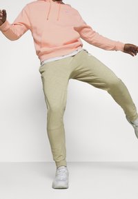 Person som bär en ljusrosa huvtröja, beige joggers och vita sneakers, mitt i ett steg med utsträckta armar mot en enhetlig vit bakgrund.