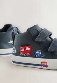 Tumši zili sporta apavi ar auduma ieliktņiem, kas attēlo transportlīdzekļus. Ar Velcro siksnām, teksturētu virsējo daļu un gumijas zoli ar baltiem un ziliem akcentiem.