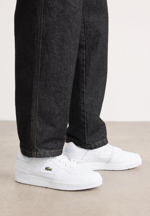 Personne portant un jean foncé et des baskets blanches Lacoste, debout sur un sol beige clair devant un fond uni.