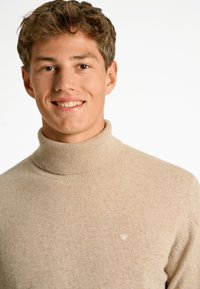 Suéter de cuello alto beige hecho de un material de punto suave, con un cuello acanalado y un pequeño logotipo bordado en el pecho.