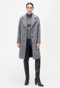 Grauer Fischgrät-Wollmantel mit Reverskragen, drei Knöpfen und Seitentaschen. Über einem grauen Pullover und dunklen Jeans getragen, kombiniert mit kniehohen Stiefeln.