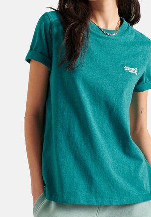 Donna che indossa una maglietta azzurra con maniche arrotolate, con un piccolo logo bianco sul petto e pantaloni chiari, accessoriate con una collana di catena dorata.