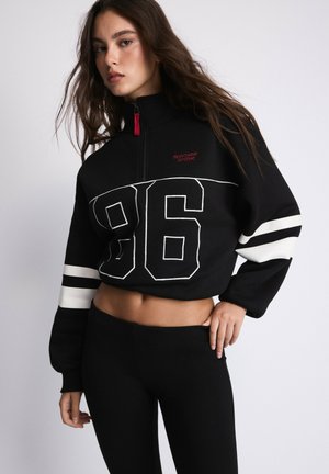 Jeune femme aux longs cheveux portant un sweat-shirt court noir avec le numéro 96 et des bandes blanches sur les manches, associé à des leggings noirs.