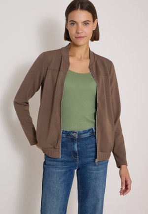 Femme portant une veste marron zippée sur un débardeur vert clair et un jean bleu, posant avec une main sur la hanche.