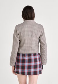 Veste brune en pied-de-poule courte avec des manches longues, associée à une jupe écossaise noire et rouge avec des accents bleu et blanc.