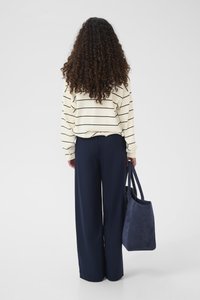 Camiseta de manga larga a rayas crema, pantalones anchos azul marino, llevando un bolso tote de ante azul marino. Rizos con textura visibles, de espaldas al espectador.