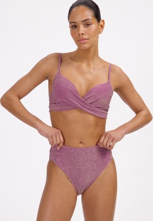 Femme portant un maillot de bain deux-pièces rose scintillant, ajustant la culotte taille haute sur un fond blanc uni.