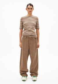 Persoon in een beige gestreepte korte mouwen shirt, losse bruine corduroy broek en witte sneakers met groene accenten, staand tegen een witte achtergrond.