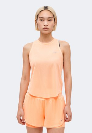 Jeune femme aux cheveux blonds courts portant un débardeur de sport pêche et un short assorti, debout devant un fond clair uni.