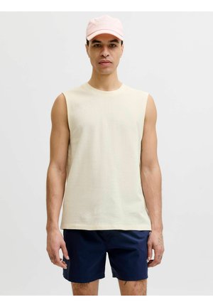 Mees kannab hele beežika värvi varrukateta särki, tumesiniseid pükse ja roosat baseballmütsi, seisab ühevärvilise heleda tausta ees.