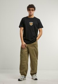 Tricou negru din bumbac cu grafică "DETROIT" și tigru, asortat cu pantaloni cargo khaki cu cusături vizibile, purtat cu adidași argintii.