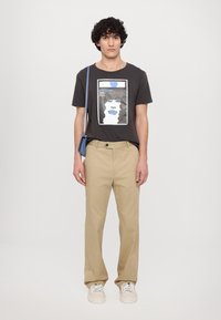 Donkergrijze graphic t-shirt met een geprint illustratie van een gezicht met krullend haar en blauwe accenten. Gecombineerd met beige broeken en sneakers.