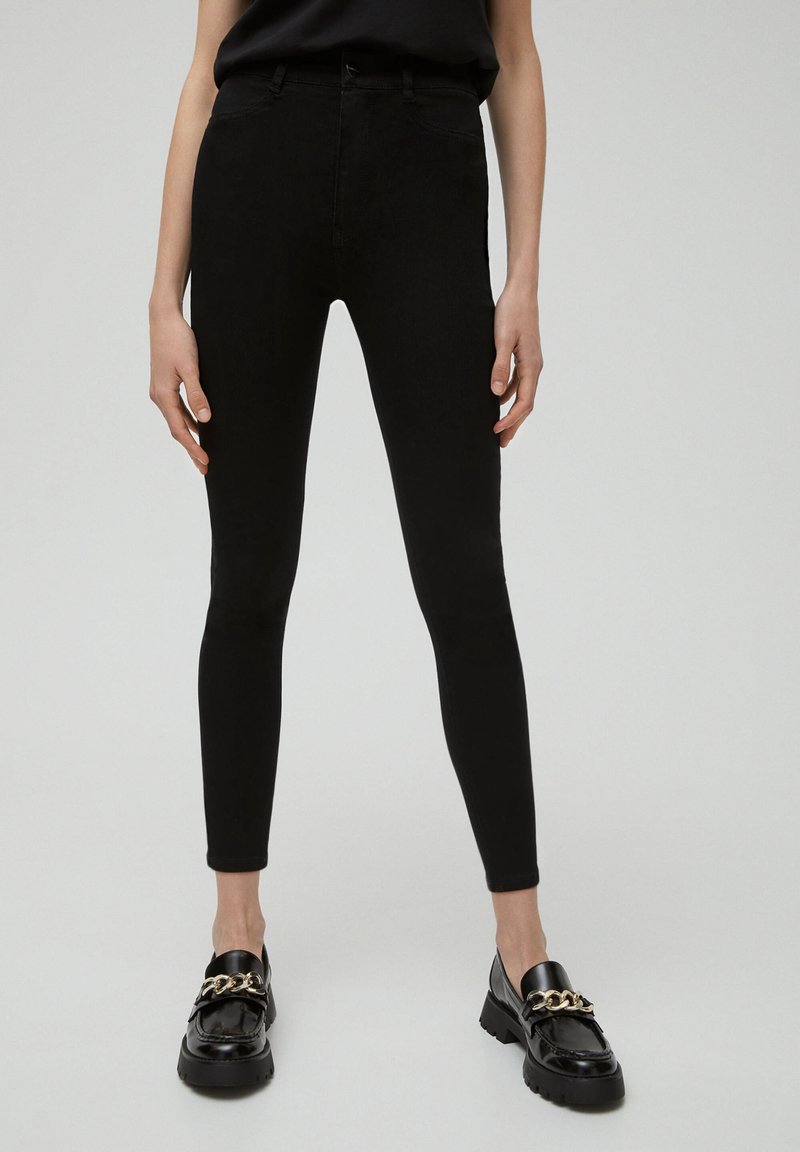 Black skinny jeans with a high waist, featuring a smooth texture, five-pocket design, and paired with glossy black loafers with gold chain detail.