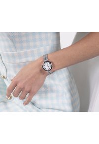 Orologio da polso argentato con quadrante rotondo bianco, indicatori delle ore neri e cinturino in maglia argentata testurizzata. La mano poggia su un tessuto a quadri blu chiaro.