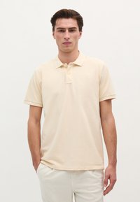 Lys beige polo skjorte i åndbart bomuld. Har en placket med tre knapper, korte ærmer og et diskret logo broderet på brystet.