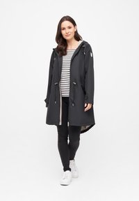 Impermeable negro con capucha, ceñido en la cintura, con un forro beige y detalles de cordones. Combinado con una blusa de rayas.