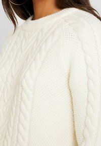 Pull crème en grosse maille avec encolure ronde côtelée et motifs torsadés texturés sur les côtés, présentant une texture douce et chaude.