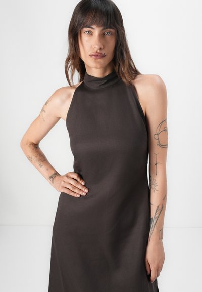 Bruine halterjurk van gladde stof, met een getailleerde top en een vloeiende rok. Tatoeage-details zichtbaar op de armen.