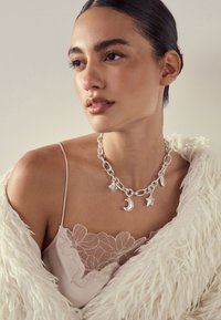 Woman with slicked-back hair wearing a white fluffy jacket, floral lace camisole, and chunky silver charm necklace with moon and star pendants.