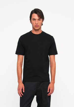 Young man with medium-length hair wearing a plain black short-sleeve t-shirt and black pants against a white background.