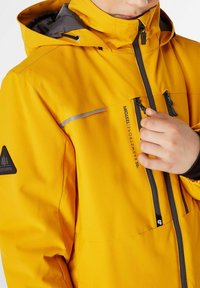 Giacca impermeabile gialla con cappuccio foderato in grigio, zip nere e dettagli riflettenti. Presenta una patch con logo sulla manica e tasche con zip.