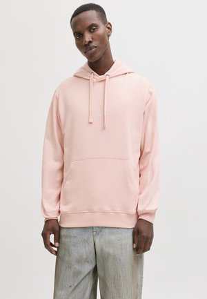 Muškarac u svijetloružičastoj majici s kapuljačom i širokim svijetloplavim trapericama stoji na jednostavnoj bijeloj pozadini.