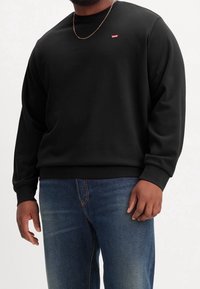 Felpa nera con collo a giro, polsini e orlo a costine, caratterizzata da un piccolo logo rosso di Levi’s sul petto. Indossata con jeans blu.