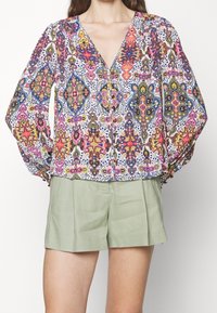 Multikleurige blouse met een patroon, lange puffmouwen, een v-hals, knopen aan de voorkant en een lichte, vloeiende stof. Gekoepeld met groene shorts.