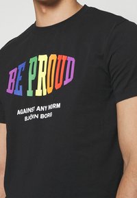 Czarny bawełniany T-shirt z wielokolorowym napisem "BE PROUD" nad białym napisem "AGAINST ANY NORM" oraz "BJÖRN BORG". Krótkie rękawy, okrągły dekolt.