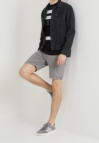 Mustat farkkutakki, musta graafinen t-paita valkoisilla raidoilla, harmaat shortsit, harmaat lenkkarit valkoisilla yksityiskohdilla, seisomassa yksinkertaista taustaa vasten.