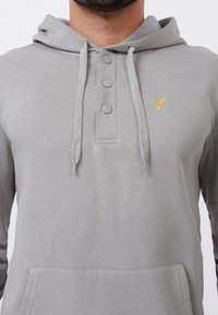 Man wearing light gray hoodie with three gray buttons, drawstrings, front pocket, and small yellow bird embroidered on the chest.