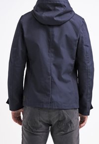 Giacca a cappuccio blu navy con una texture liscia, design aderente e dettagli delle cuciture; presenta un anello sul retro ed è abbinata a pantaloni grigi.
