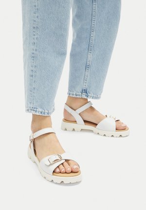 Sandalias blancas de cuero con suela gruesa, que incluyen una hebilla frontal y una correa en el tobillo. Combinadas con pantalones vaqueros cortos de mezclilla azul claro.